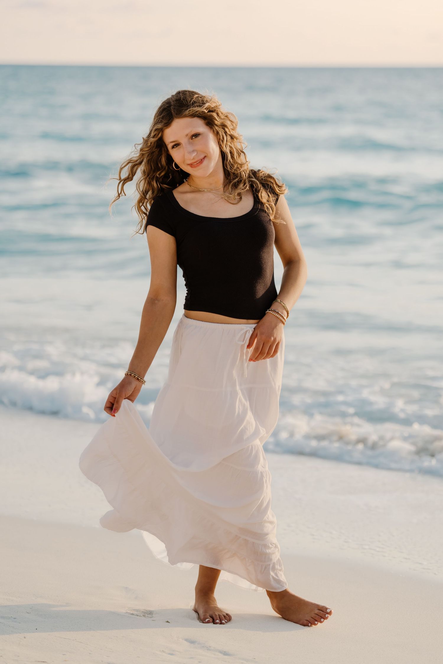 Barefoot woman in black crop top and flowing white skirt poses on beach at sunset with waves in background. Senior photos grand cayman