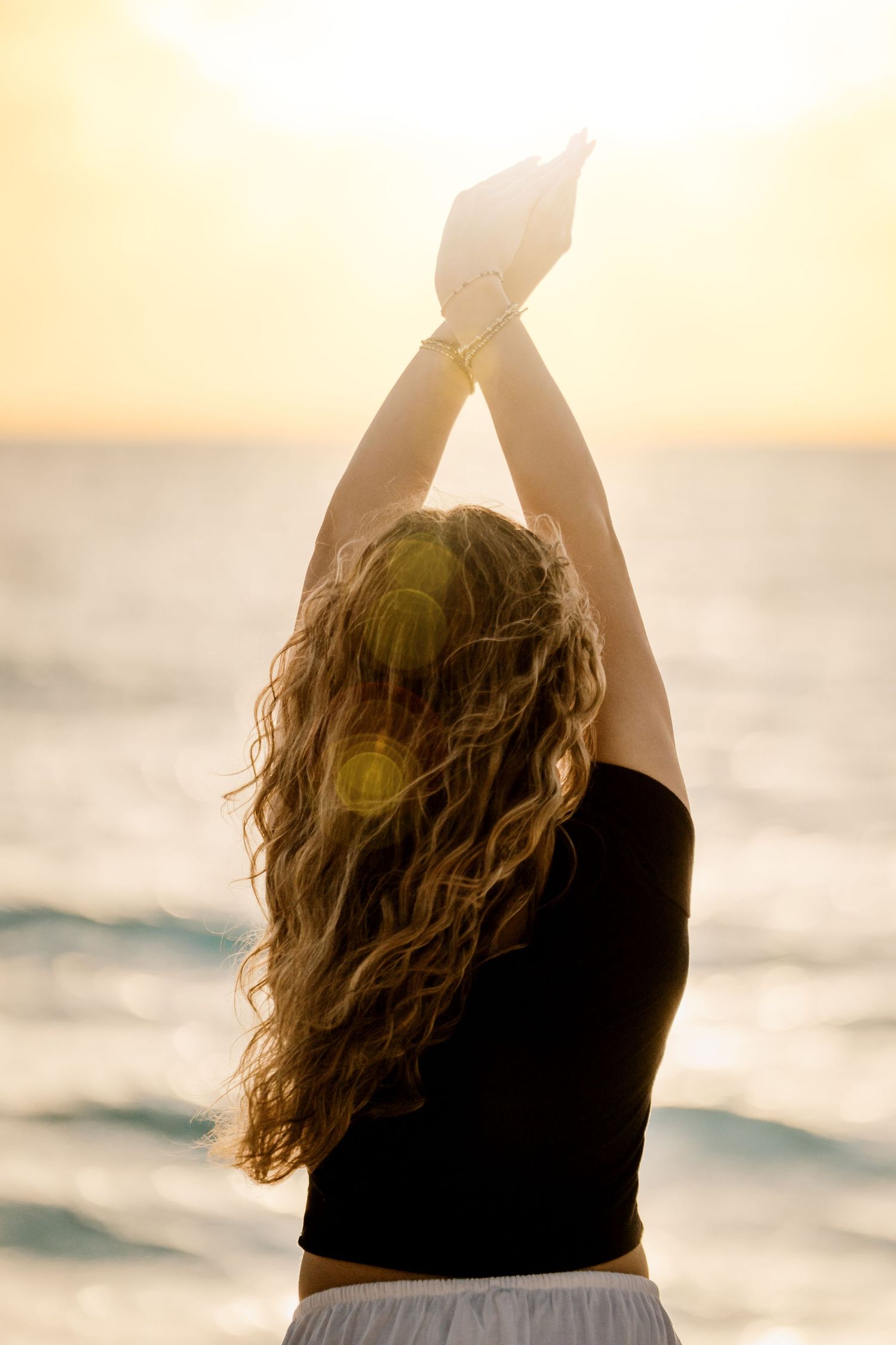 Silhouetted figure with long wavy hair raises arms toward golden sunset over ocean waves in backlit warm glow. Senior Photos