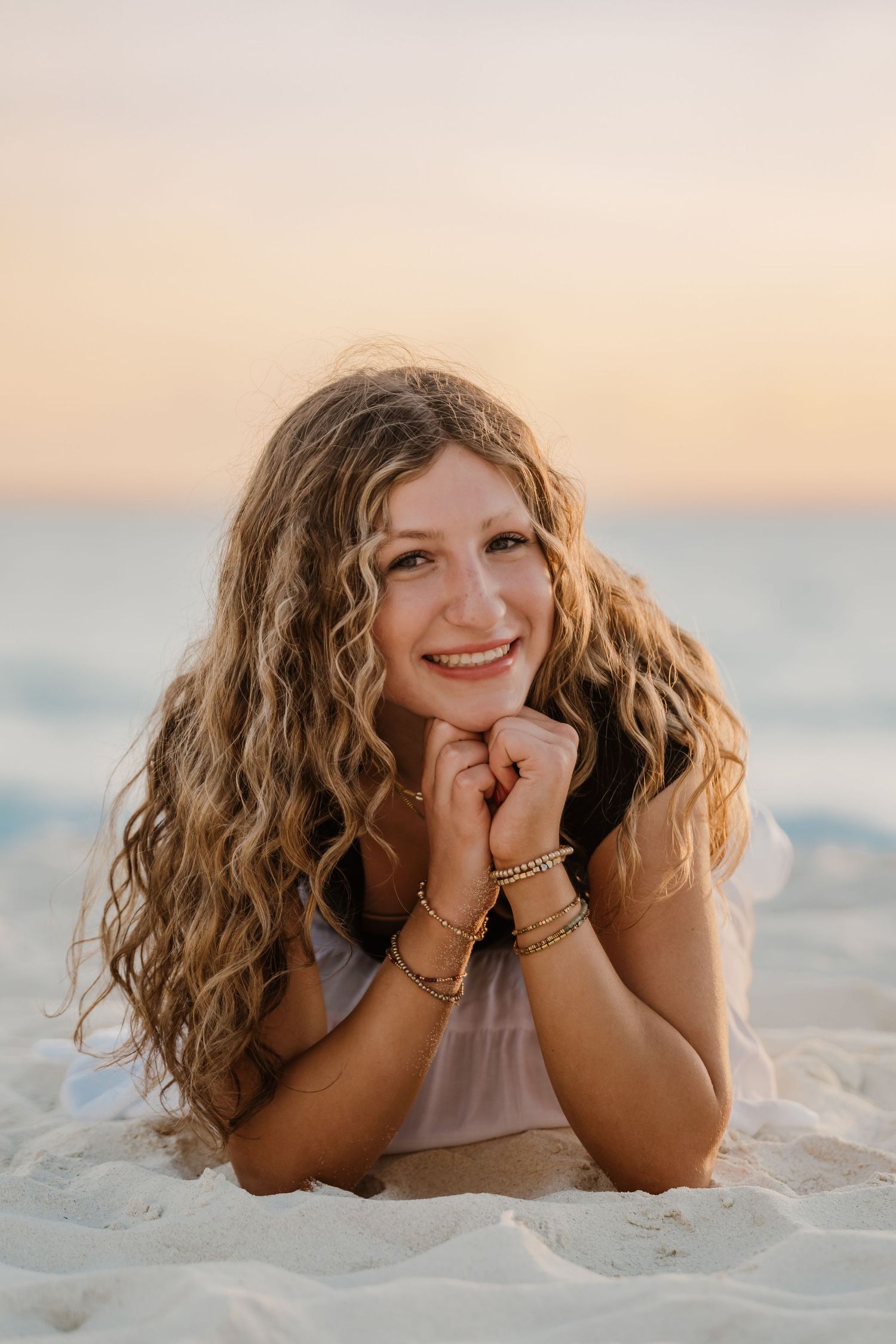 A person with long curly blonde hair lies on white sand at the beach during sunset with a warm glowing sky. Senior Photos