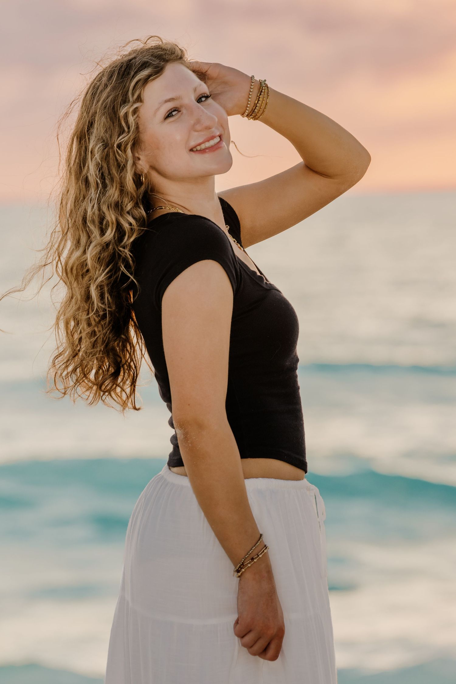 A person in a black top and white skirt poses joyfully against a sunset beach backdrop with flowing curly hair.  Senior Photos