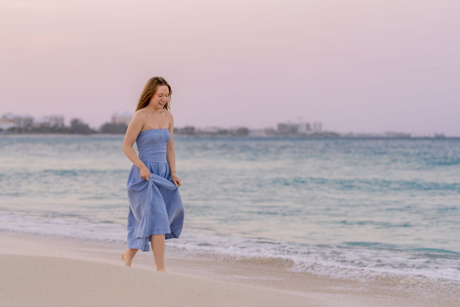 A person in a flowing blue dress walks along a sandy beach at sunset with ocean waves in the background. Senior Photos
