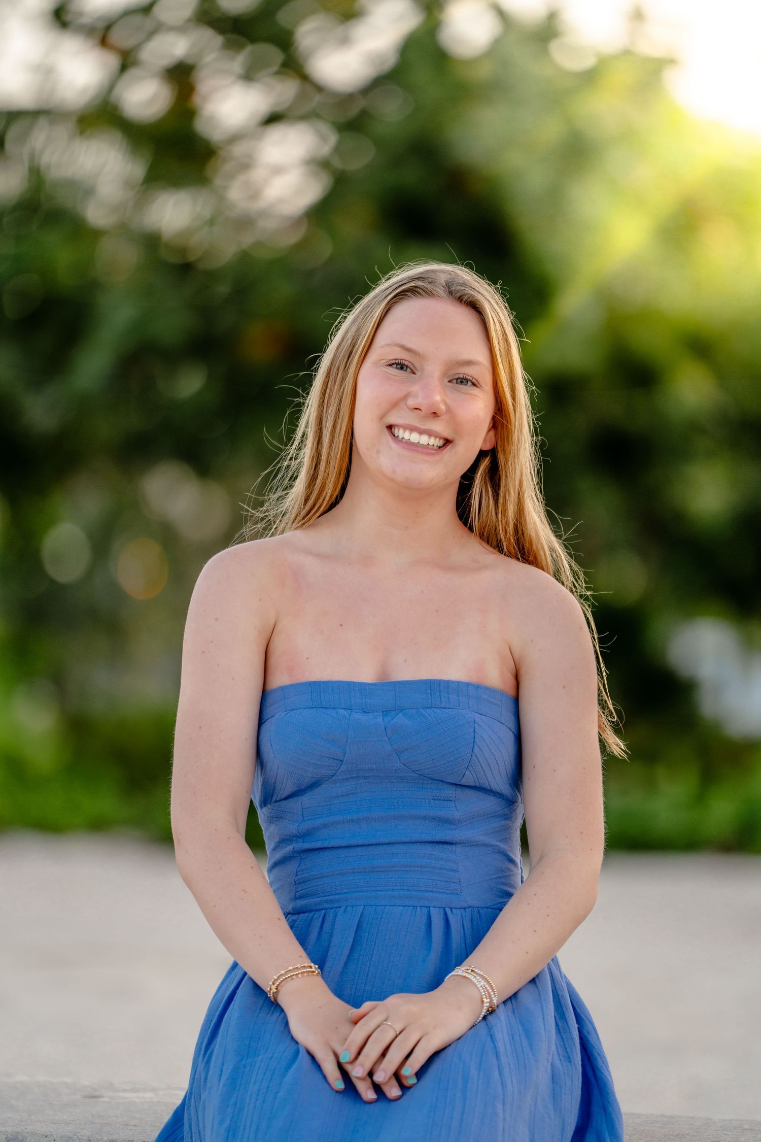 A joyful portrait of someone in a strapless blue dress with flowing blonde hair standing outdoors at golden hour. Senior Photos