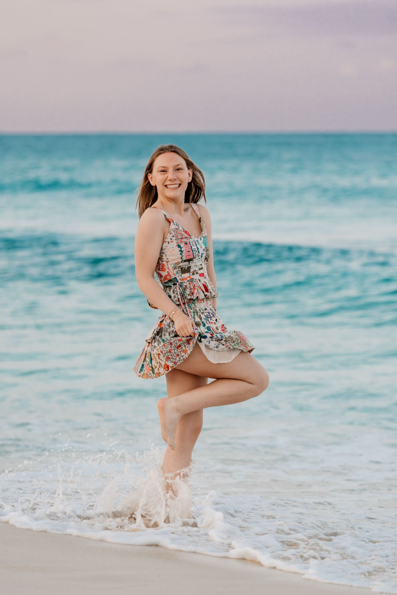 A person in a floral sundress playfully poses at the beach while waves splash around their feet at sunrise. Senior Photos