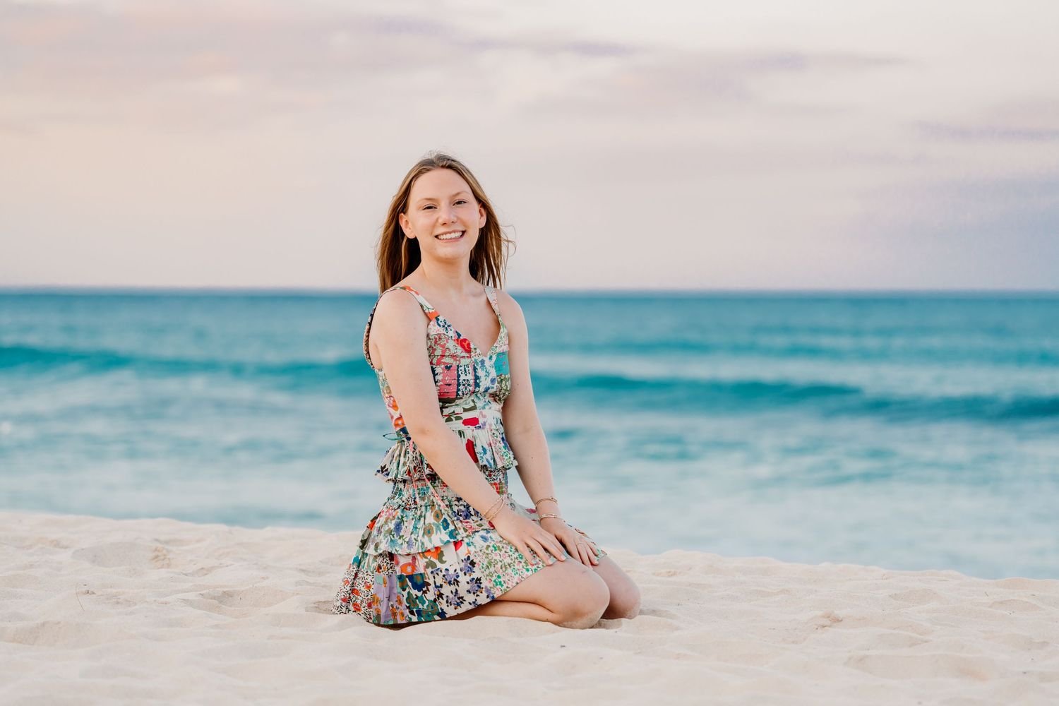 Person in a floral sundress kneeling on a tropical beach with turquoise ocean and cloudy sky in background. Senior Photos