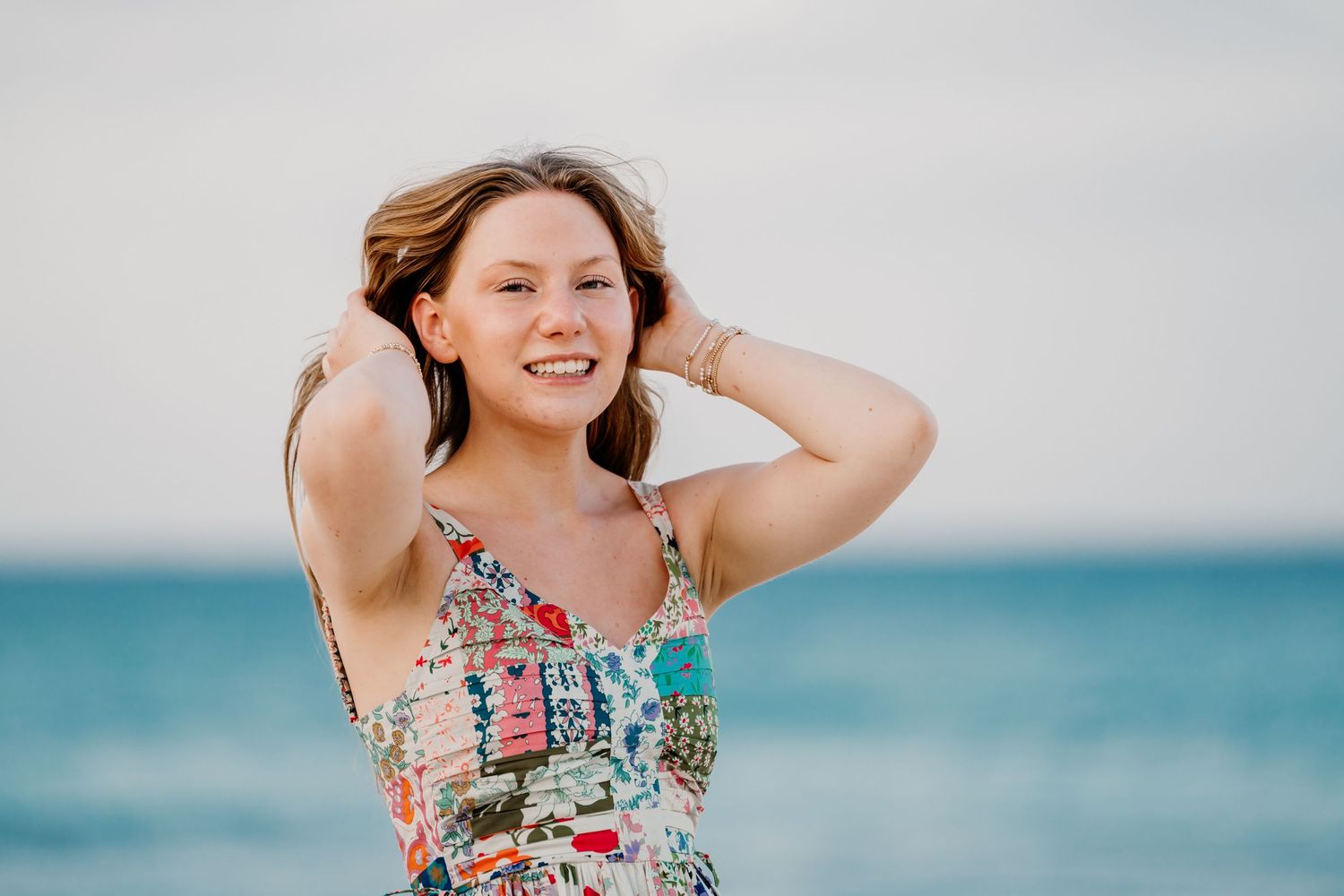 Woman in floral sundress smiles and touches her hair while standing at the beach with turquoise ocean in background. Senior Photos