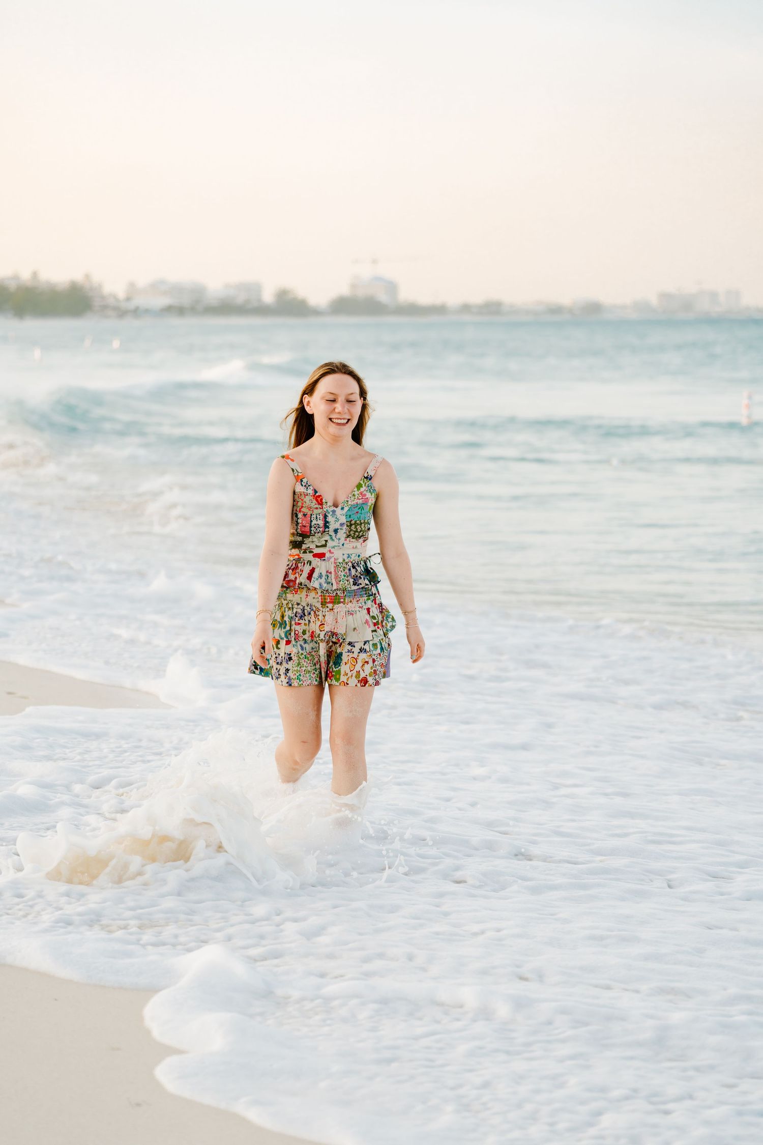 Someone wearing a colorful floral dress walks joyfully along the shoreline as waves lap at their feet during sunset.