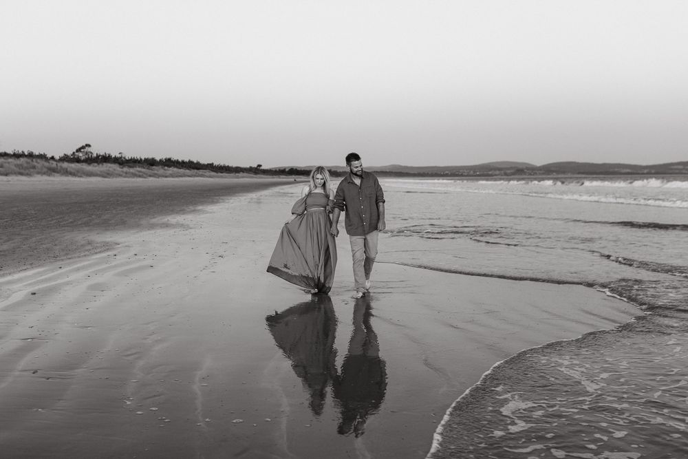 Black and white photo of a couple walking hand in hand along a reflective beach shoreline.