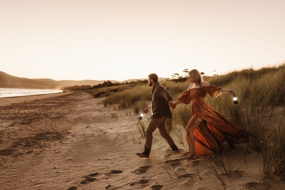 Couple walking through coastal sand dunes at golden hour, woman in elegant rust-orange gown.