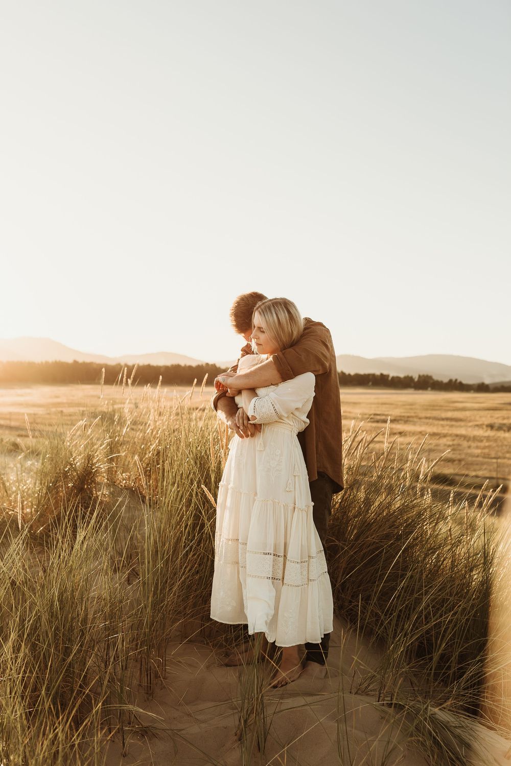 Couple embracing in golden hour light among tall grass in an open field, woman in white dress.