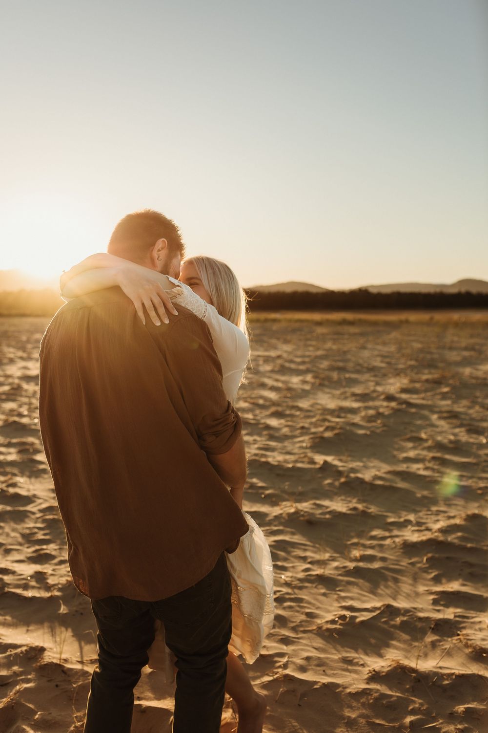 Couple sharing a tender embrace on sandy beach at sunset, man in brown shirt and woman in white dress.