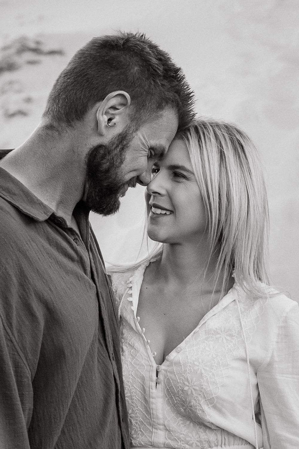 Black and white photo of a smiling couple touching foreheads intimately on a beach.