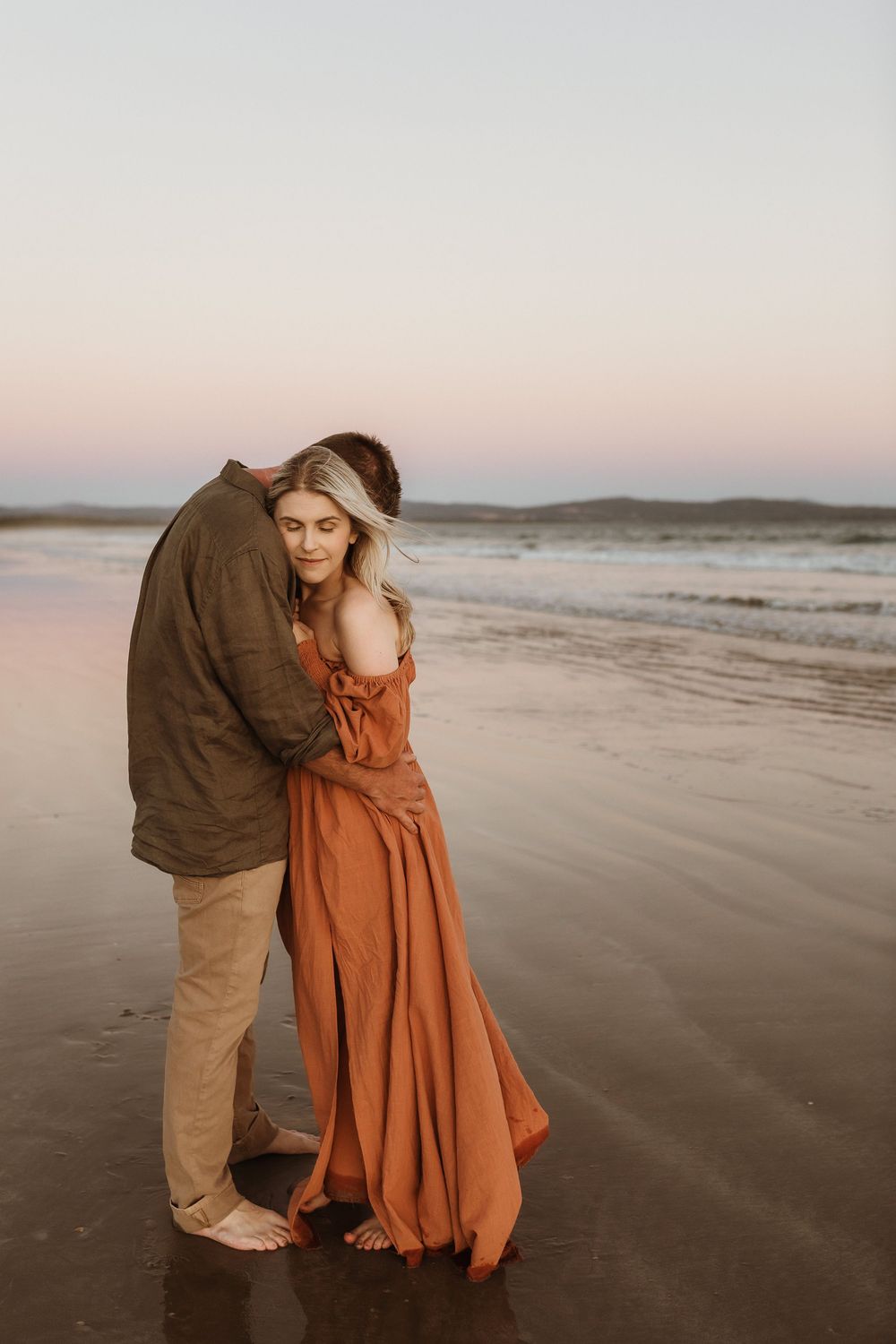 Couple embracing barefoot on a reflective beach at sunset, woman in flowing rust-orange dress.