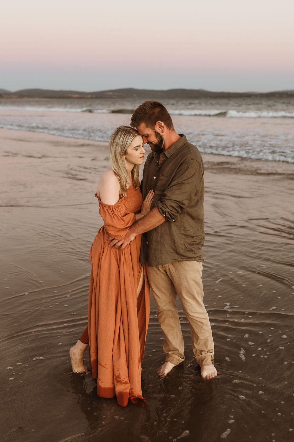 Barefoot couple standing in shallow beach water at sunset, woman in rust maxi dress embracing man.