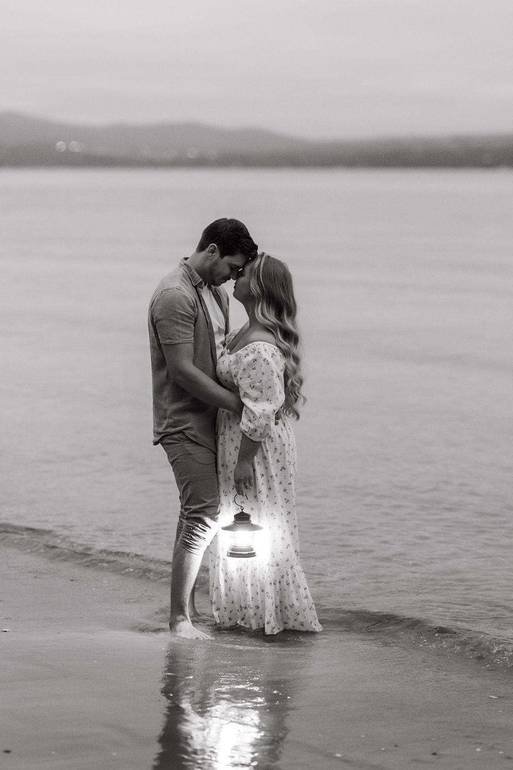 Black and white photo of a romantic couple kissing on a beach shore, holding a glowing lantern near calm water.