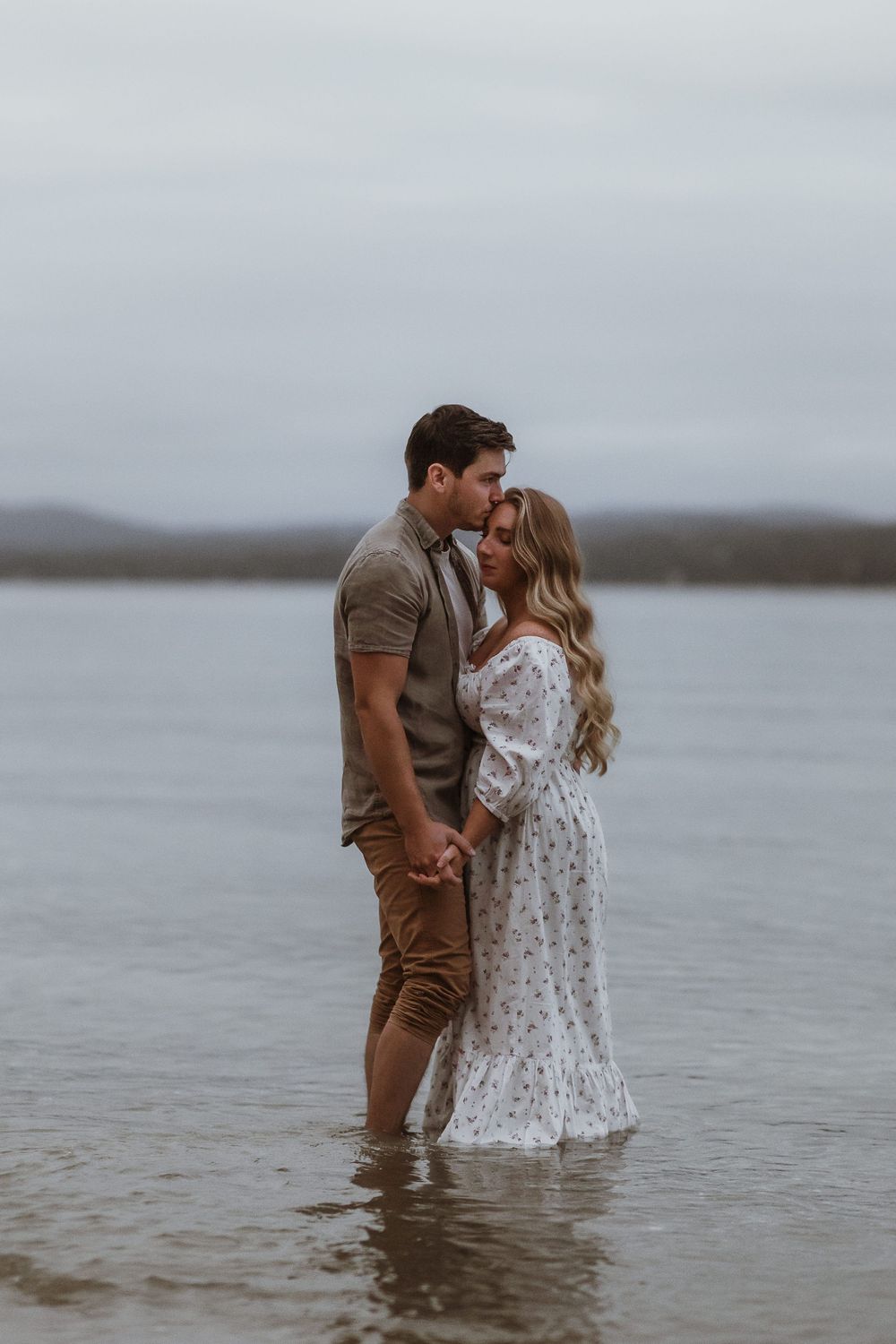 A couple shares a romantic kiss while standing in shallow water, the woman in a white floral dress and man in casual earth tones.