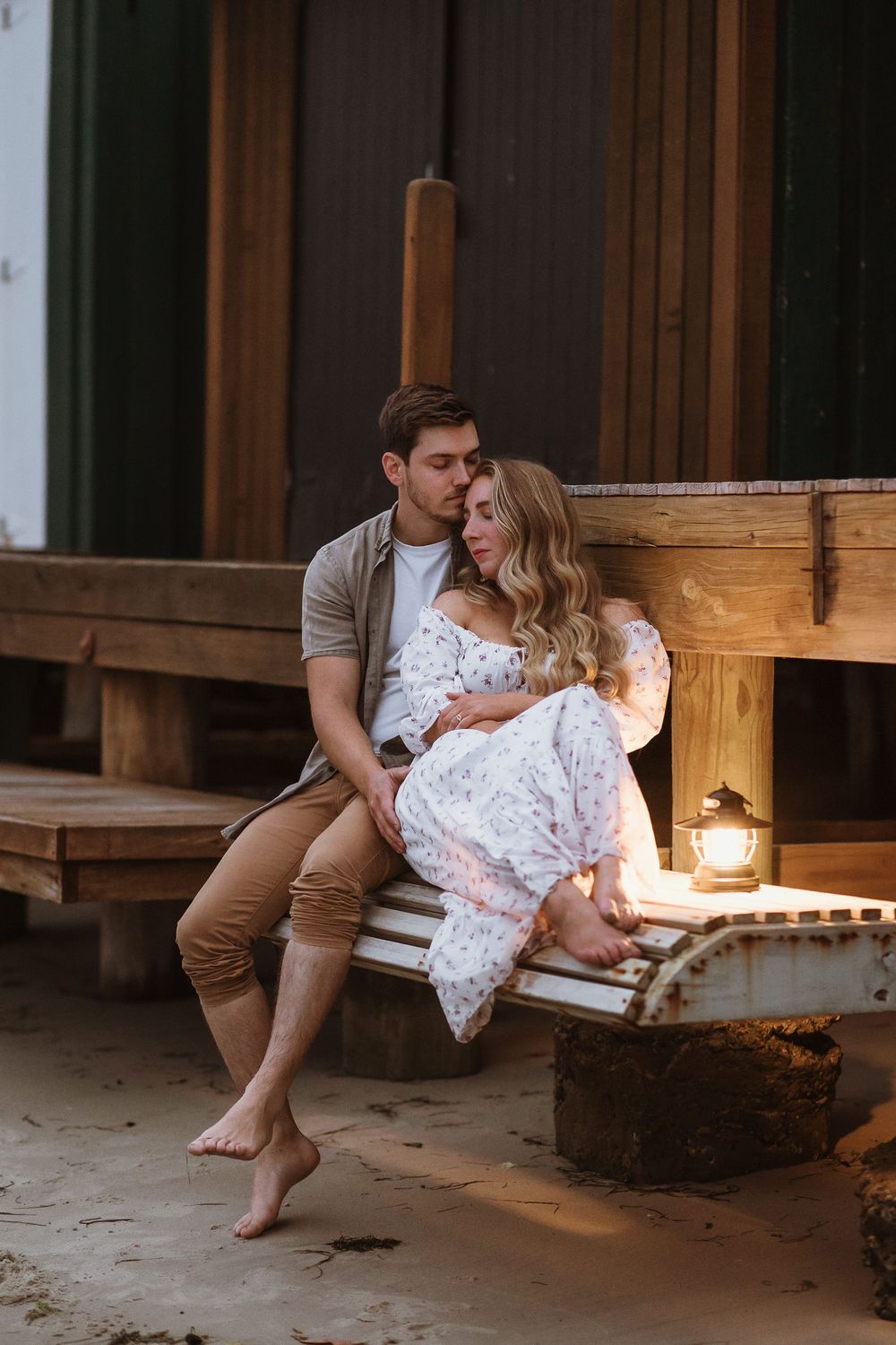Romantic couple sitting on wooden porch steps, man kissing woman's head, candlelight glowing beside them at dusk.