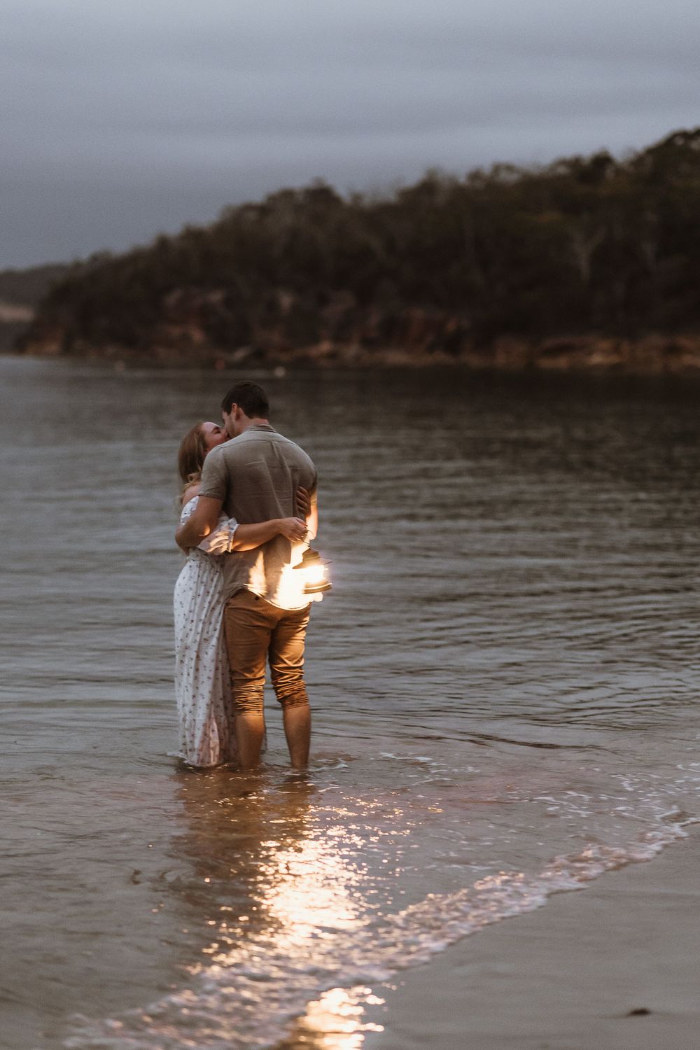 Couple embracing romantically at dusk, standing in shallow ocean water holding a glowing light on a serene beach.