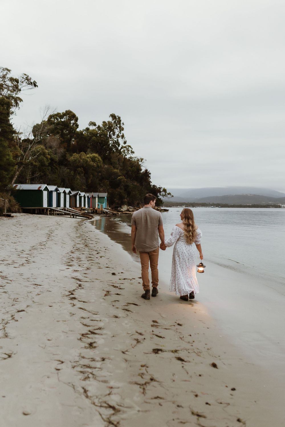 Couple walking hand in hand along a sandy beach at dusk, woman in white dress holding a lantern, beach huts in background.