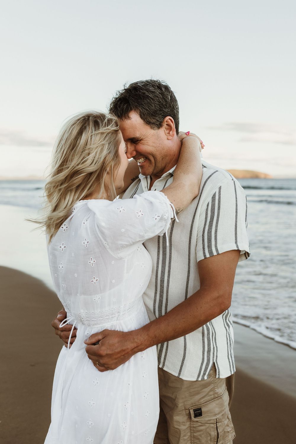A smiling couple embraces romantically on a sandy beach at sunset, woman in white dress, man in striped shirt.