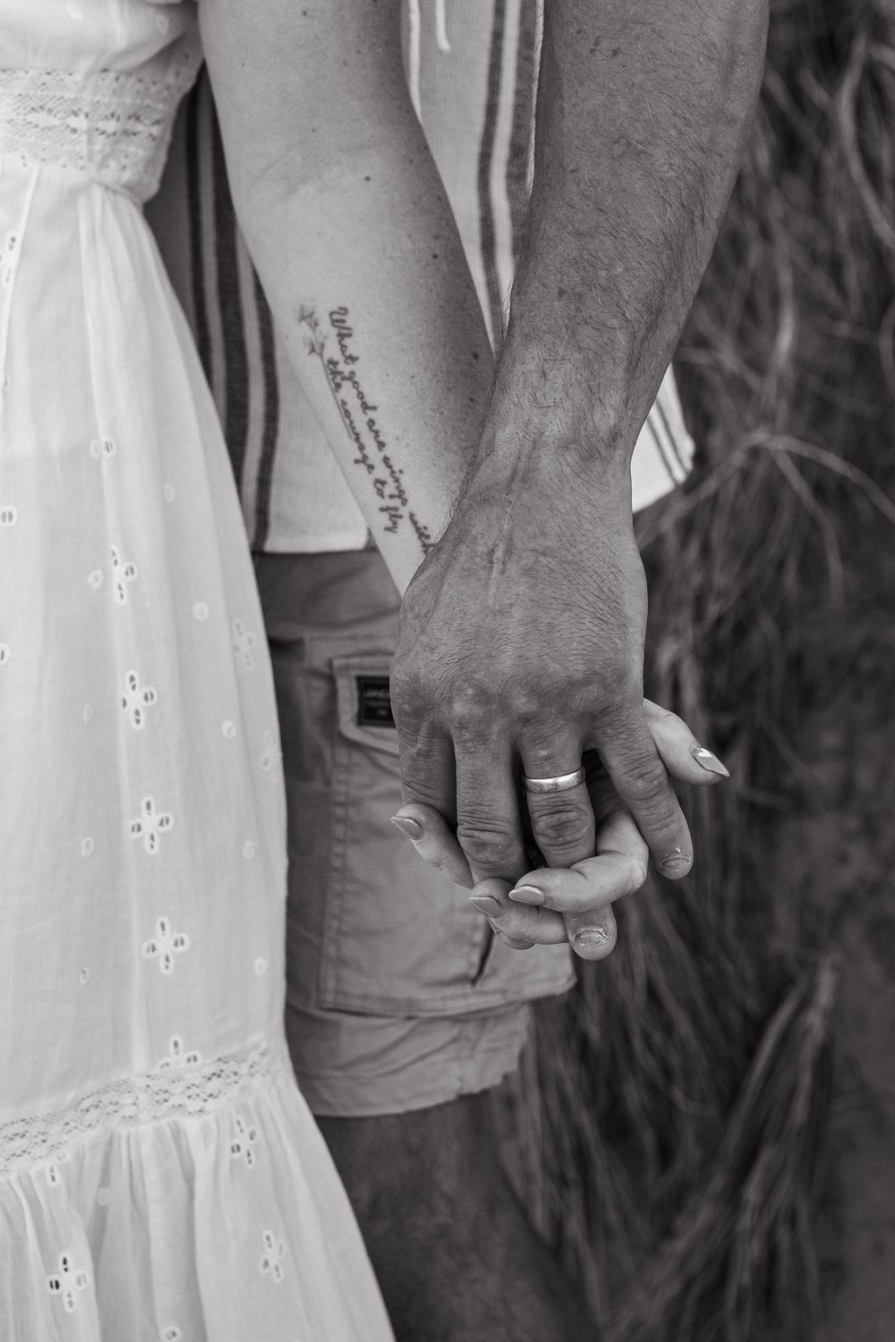 Black and white photo of a couple holding hands, showing wedding rings, tattoo, and white dress.