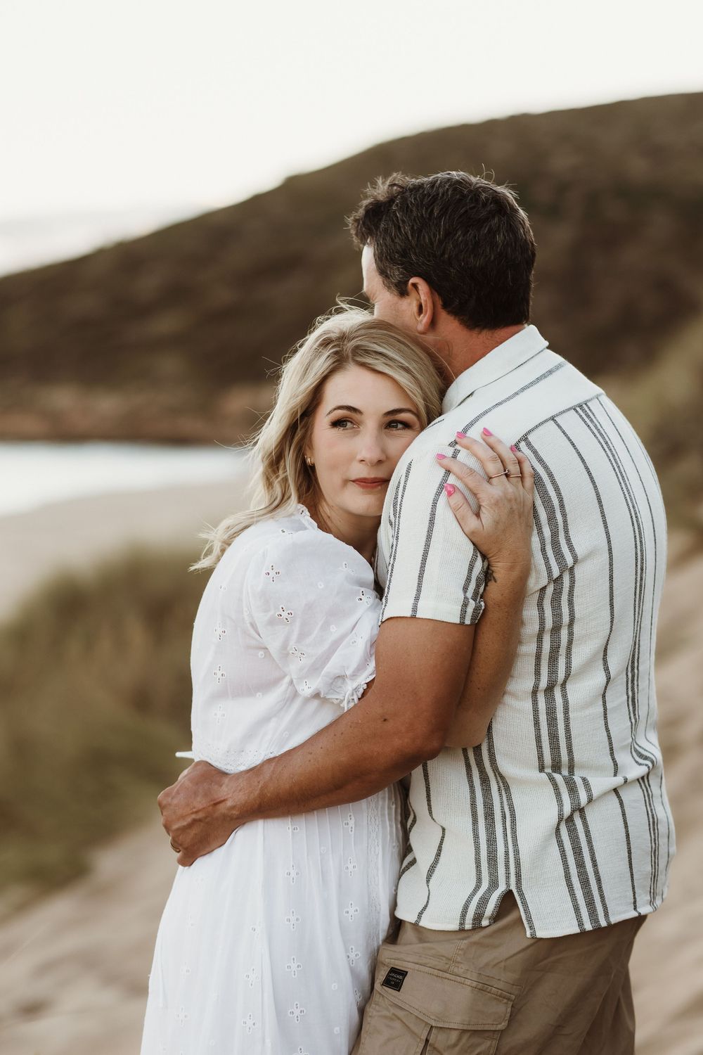 Couple embracing on a sandy beach at sunset, woman in white dress looking at camera, man in striped shirt.