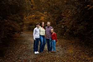 A group poses for autumn family portraits in a wooded setting with warm brown and orange tones in the background.