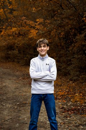 A figure in a white sweater and blue jeans stands against an autumn forest backdrop with orange foliage.