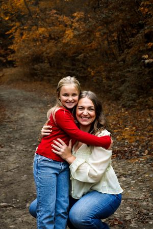 A heartwarming autumn scene shows a child in red hugging someone in a cream sweater on a leaf-covered forest path.