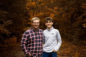 Two people pose together outdoors in autumn with vibrant orange foliage in the background.