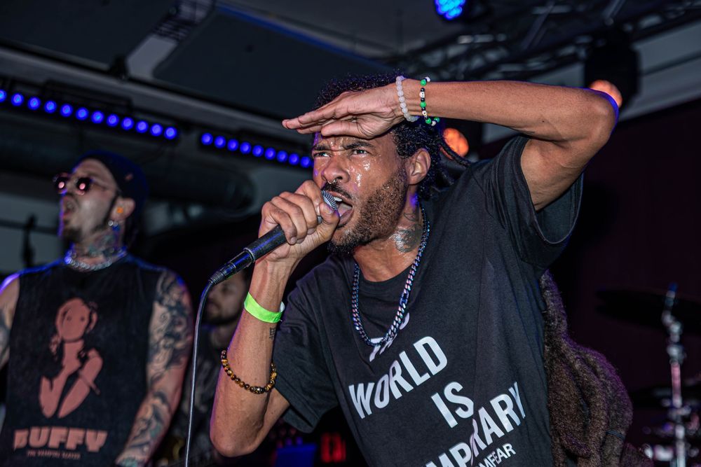 Man performing on stage with microphone, wearing black World Is Ordinary shirt, with tattooed musician in background.