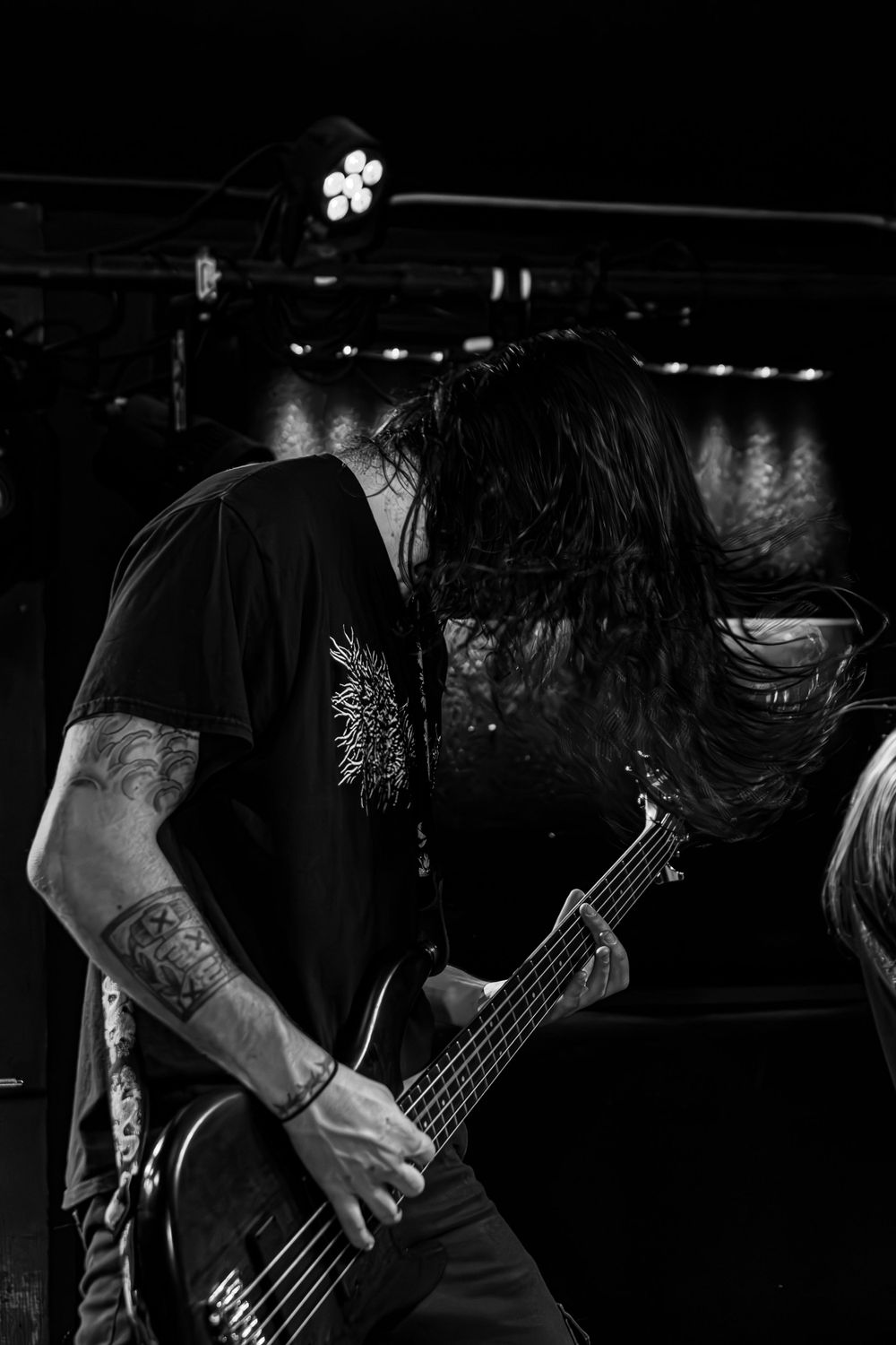 A bassist with long hair performing intensely on stage in black and white.