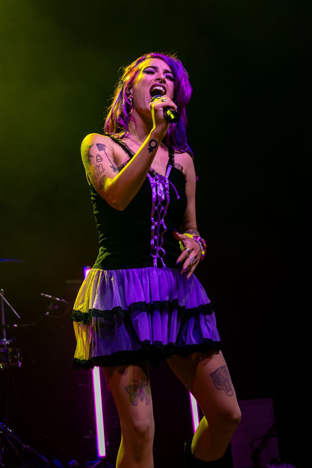 Concert performer in black and purple dress singing passionately under colorful stage lights.