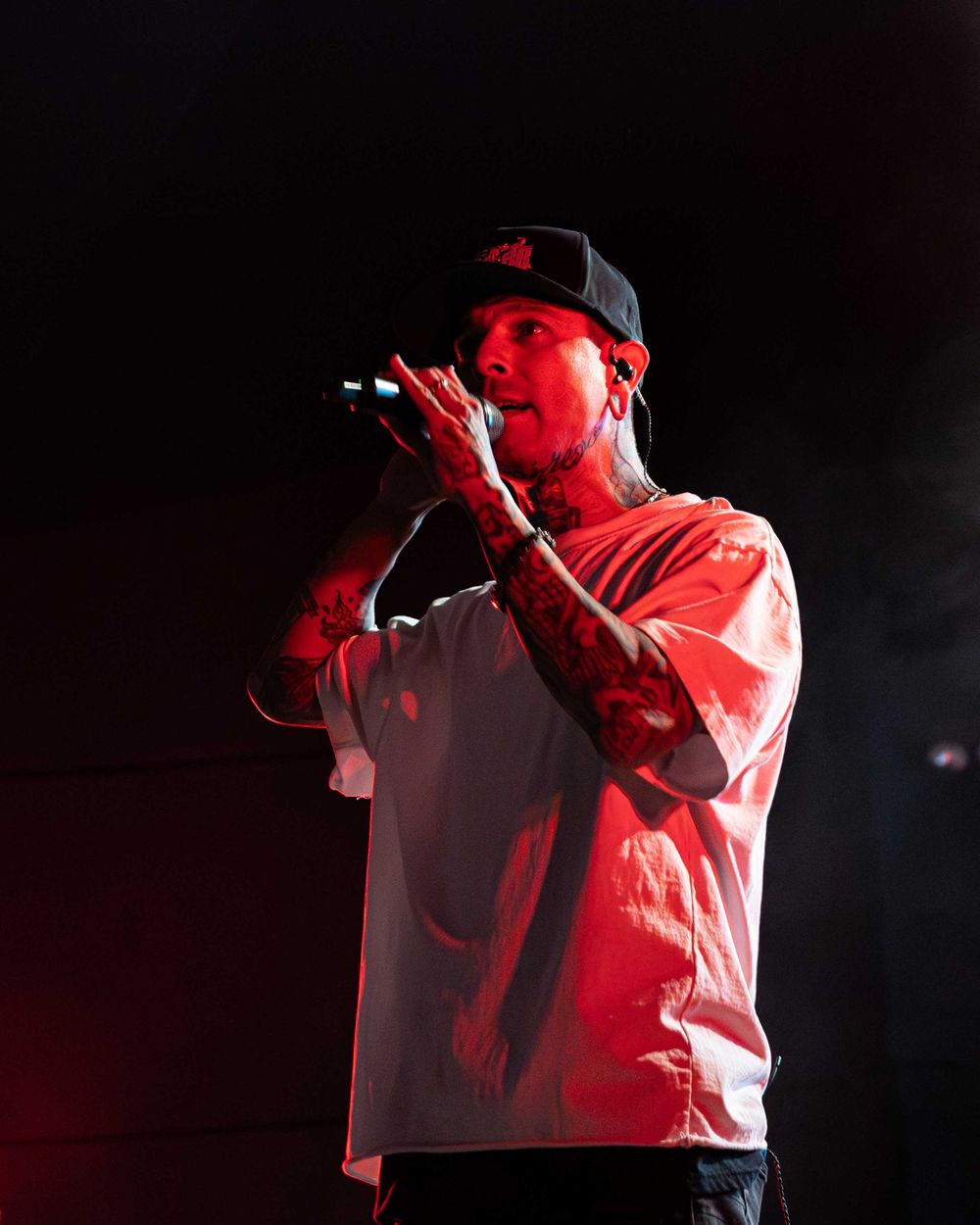 Tattooed male rapper performing on stage holding microphone under red stage lighting wearing white tee and cap.