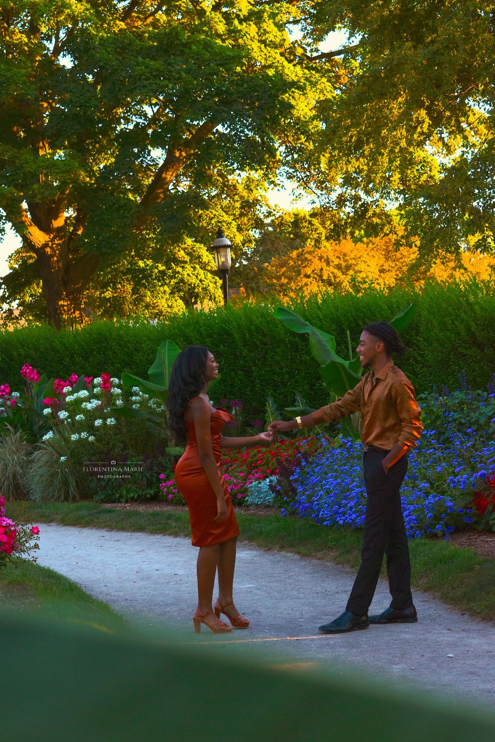 Two people converse on a garden path surrounded by blue and pink flowers at sunset.