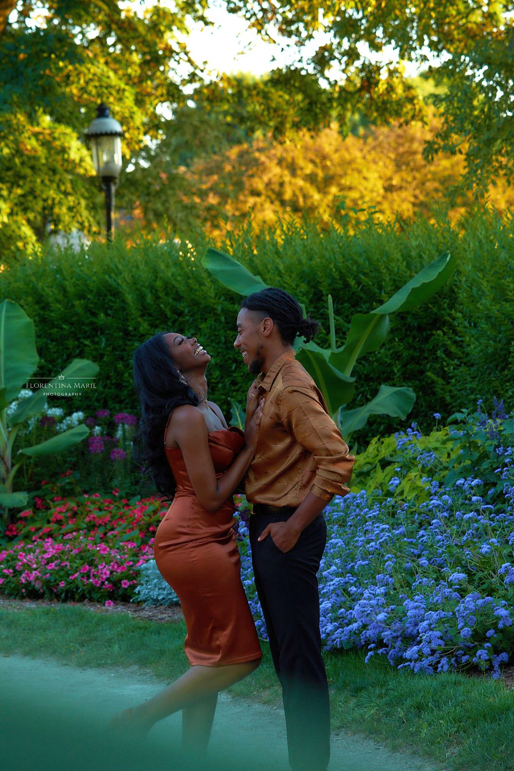 A romantic couple stands together in a lush garden with colorful hydrangeas and a lamp post at golden hour.