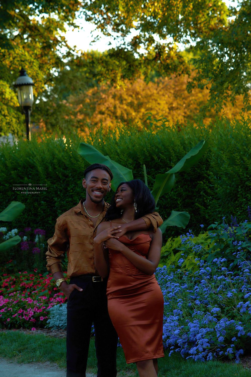 A couple shares a romantic moment in a garden with colorful flowers and lush greenery at sunset.