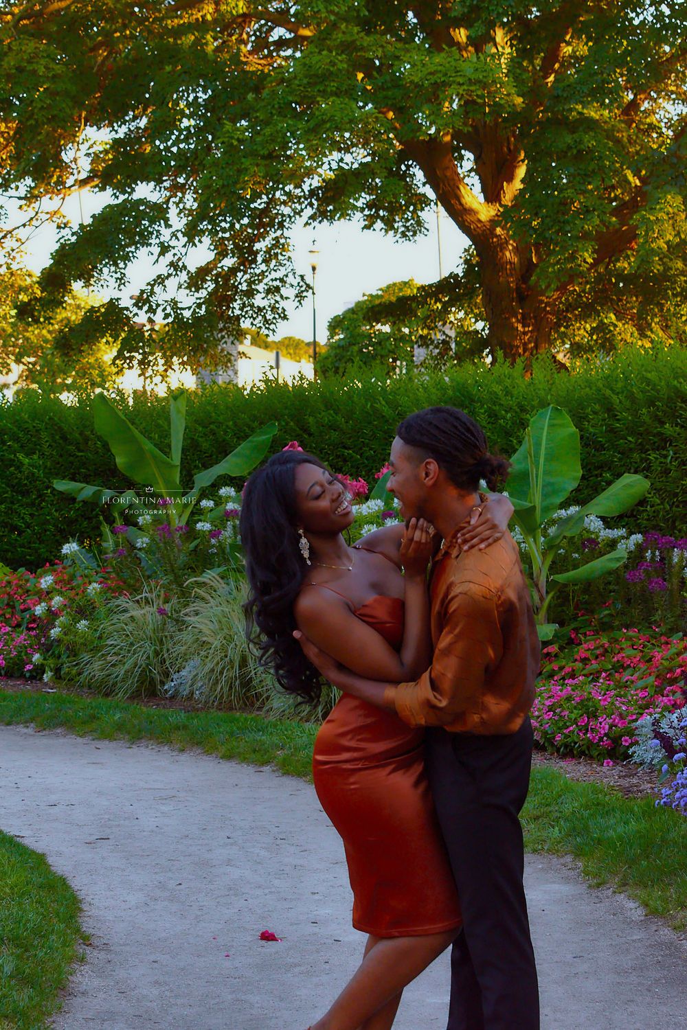A couple in coordinated orange and brown outfits dance together on a garden path at golden hour.