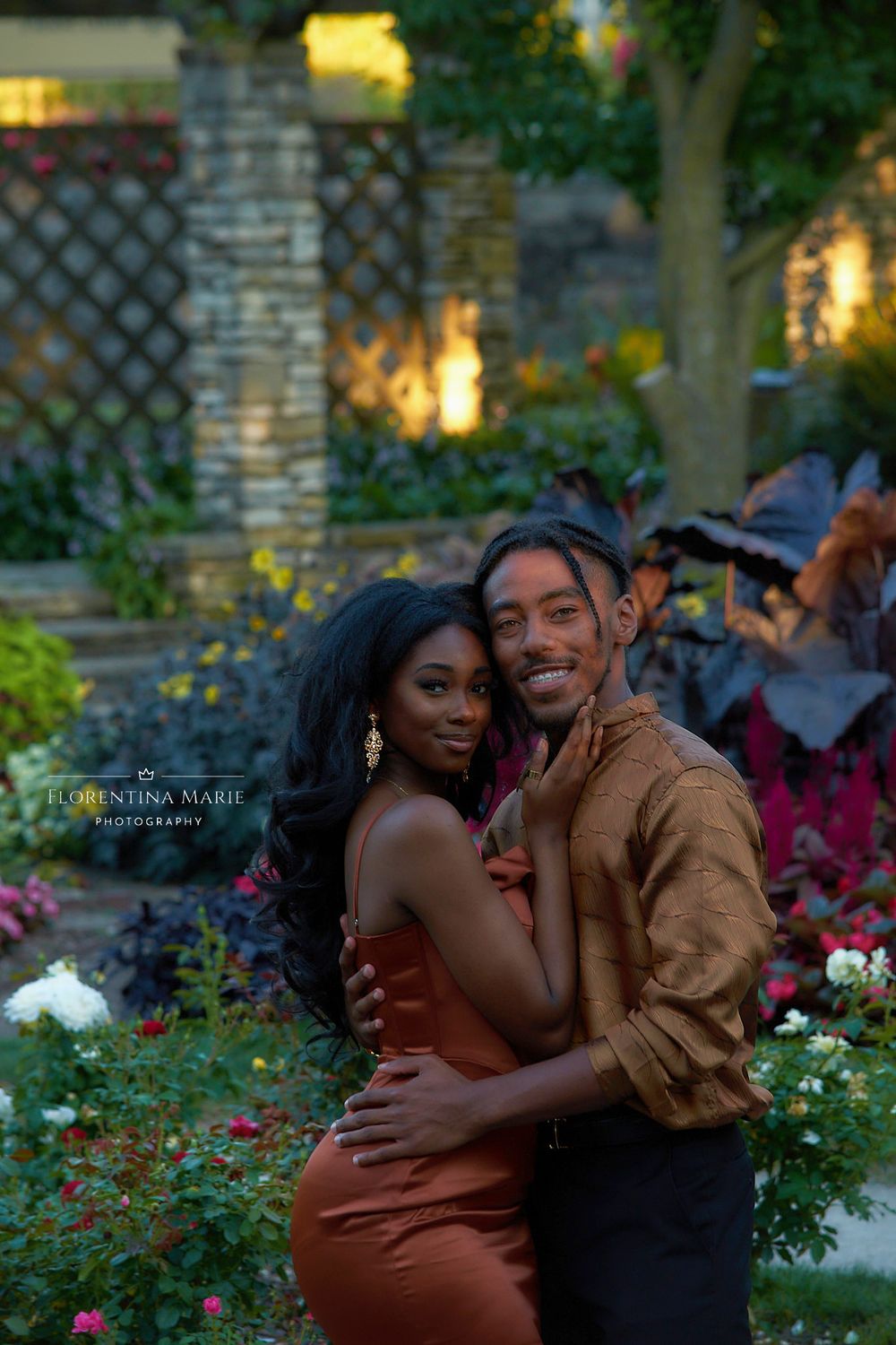 A couple in formal attire embraces in a beautiful garden setting with stone pillars and florals.