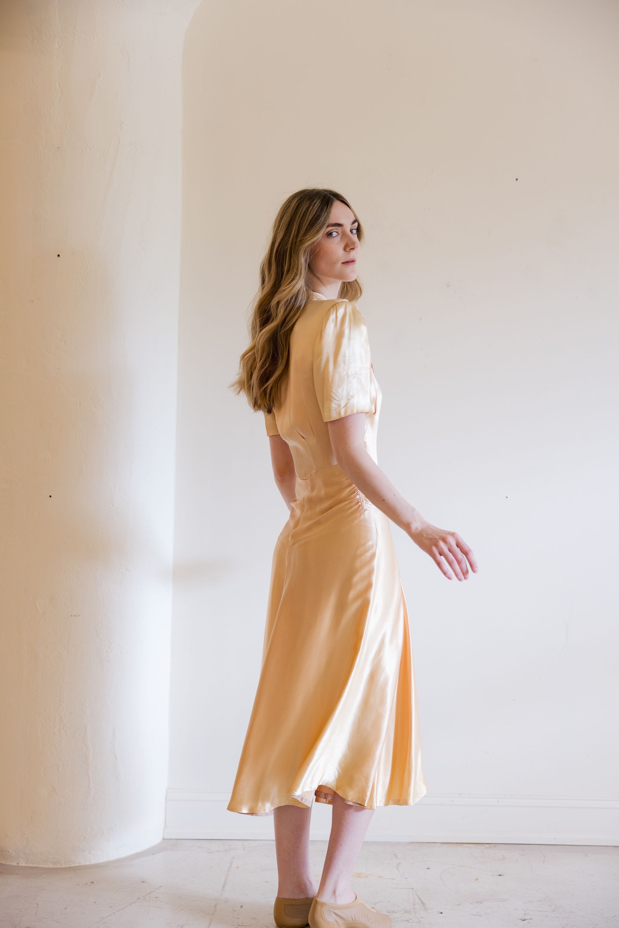 Woman in yellow satin midi skirt and cream top posing gracefully against a white wall.