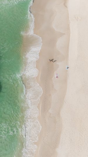 Aerial drone view of turquoise ocean waves washing onto a white sandy beach with tiny figures below.