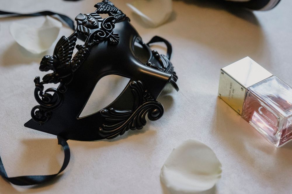 Ornate black masquerade mask with perfume bottle and wedding details on light surface.