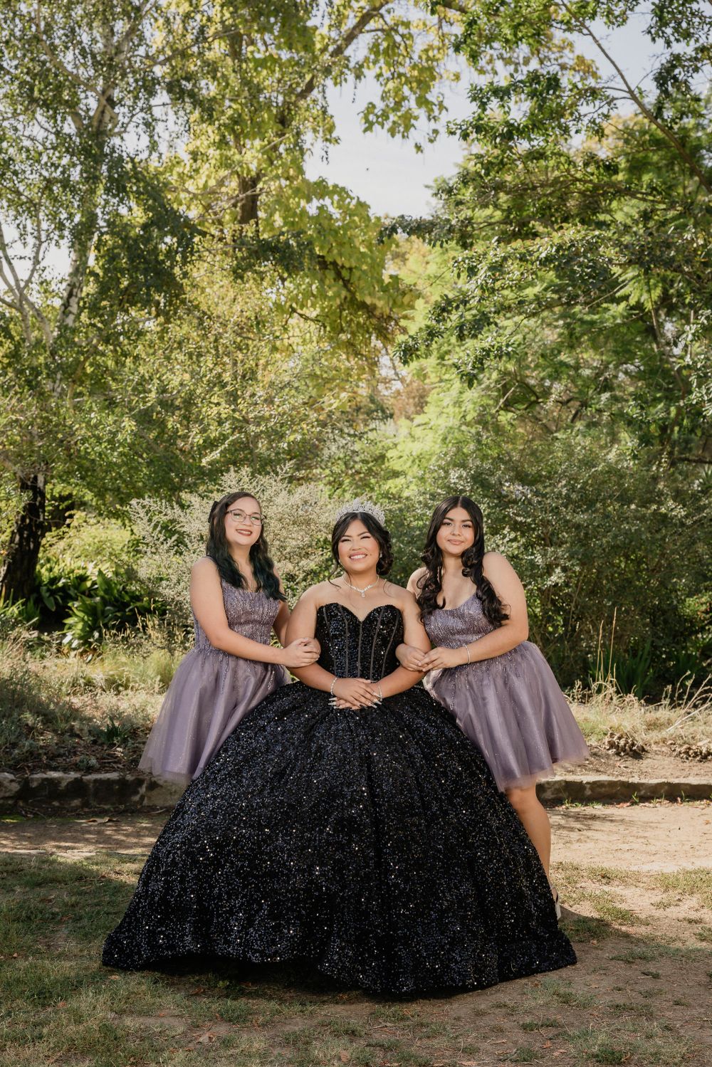 Three people posing in formal attire with a sparkly black ballgown and lavender dresses in a garden setting.