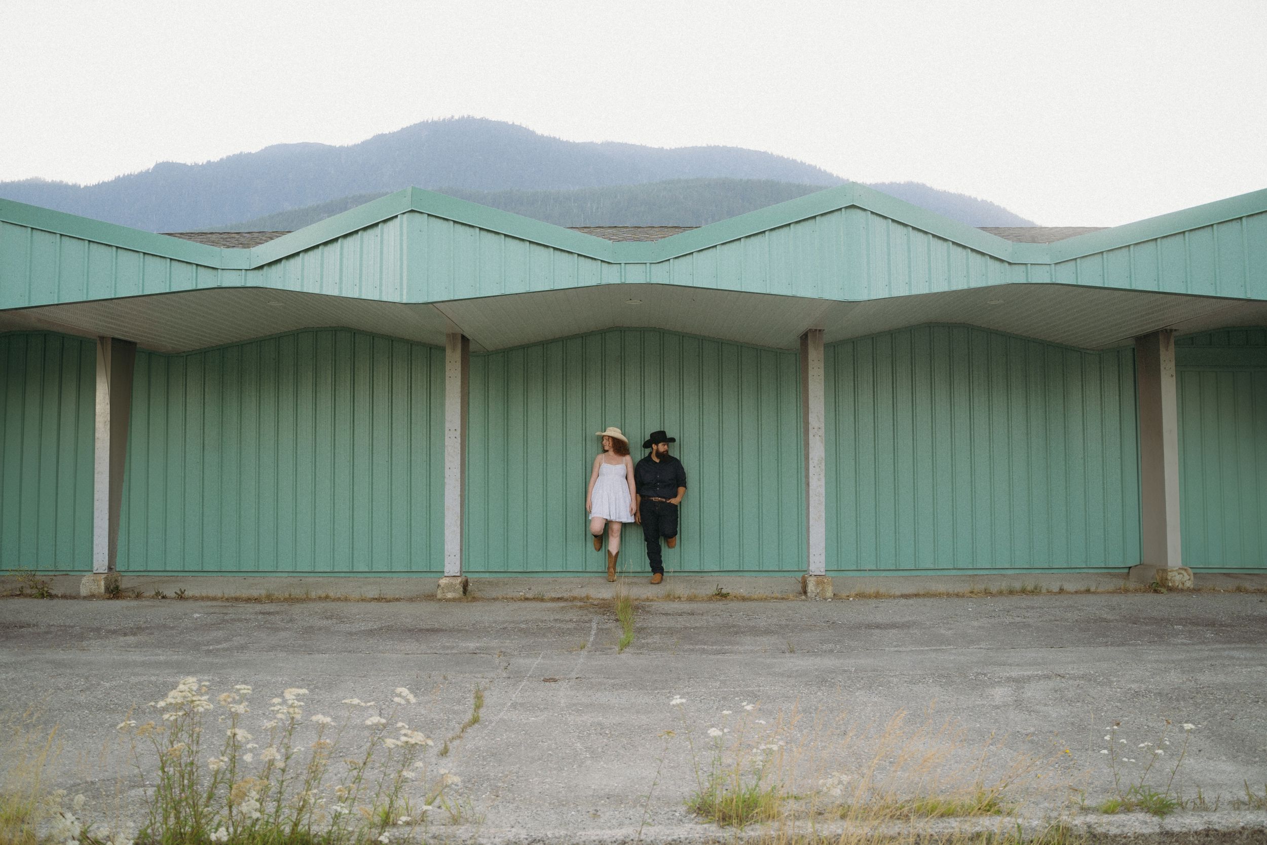 Couple standing against industrial green storage units with mountains in background.