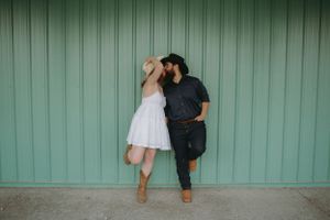 Couple embracing against turquoise industrial wall in casual attire.