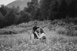 Couple shares intimate moment in wildflower field with mountains in background.