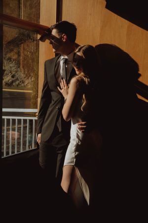 A couple in formal attire stands in dramatic lighting creating shadows against a wall.