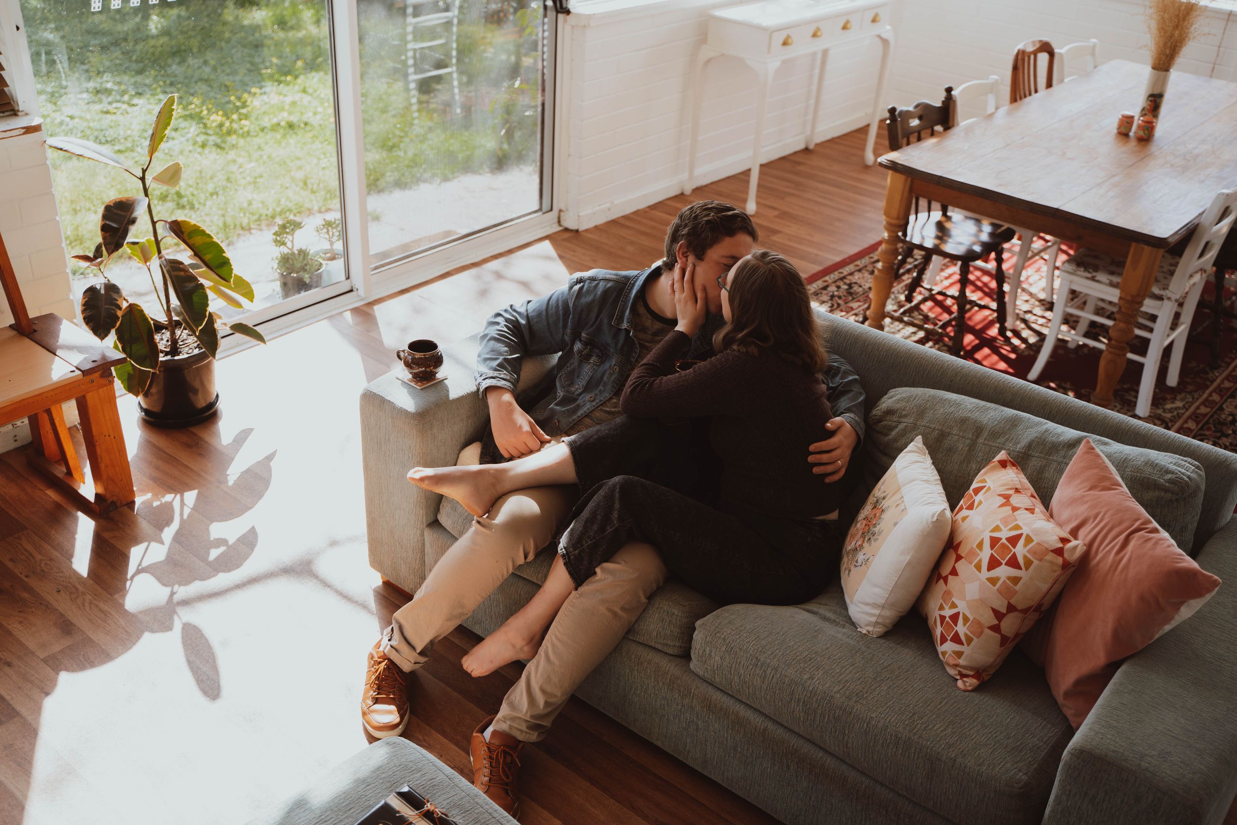 A cozy living room scene with sunlit windows, plants, and someone relaxing on a grey couch with orange throw pillows.