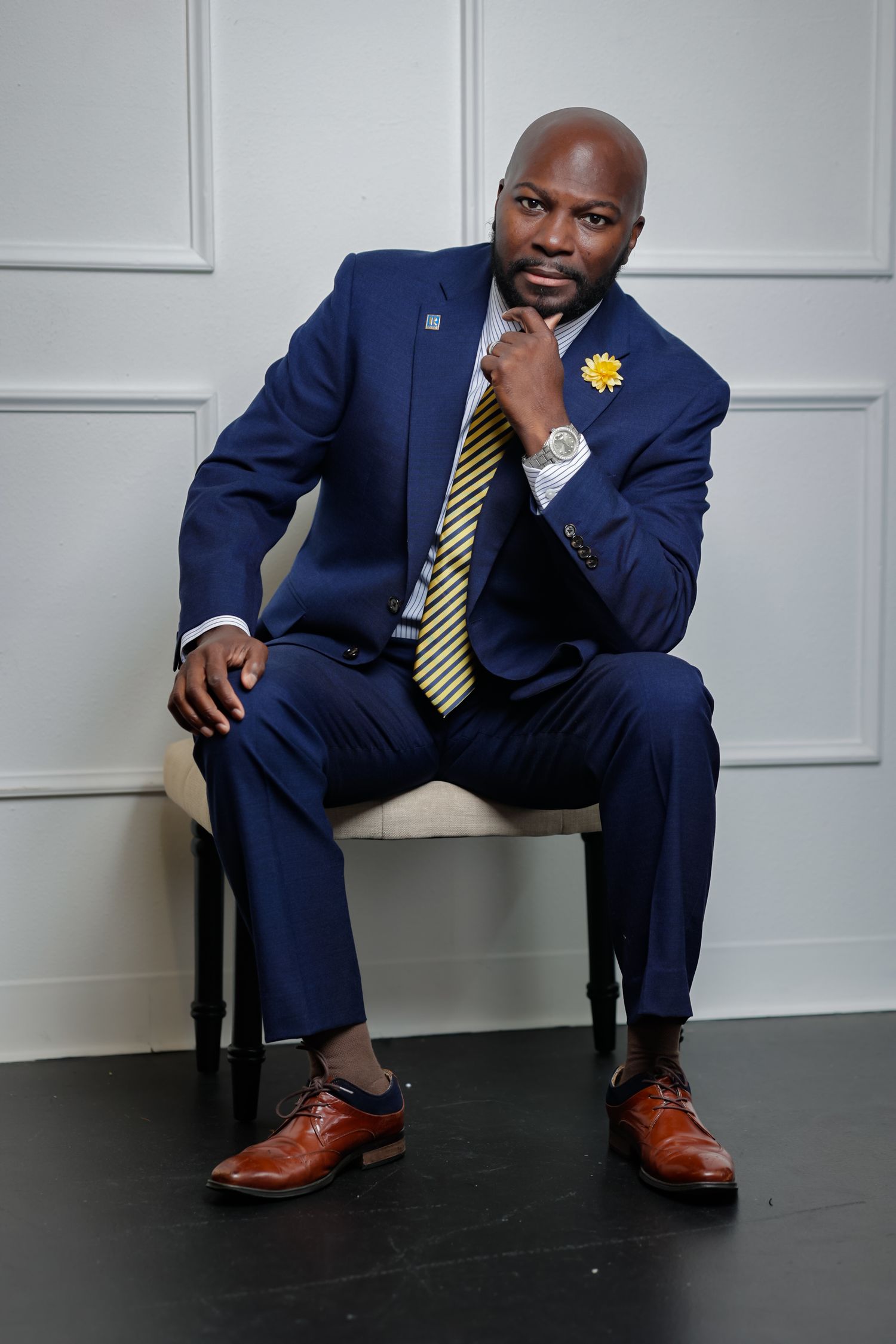 Professional Black man in navy blue suit with gold tie sitting confidently in studio portrait.