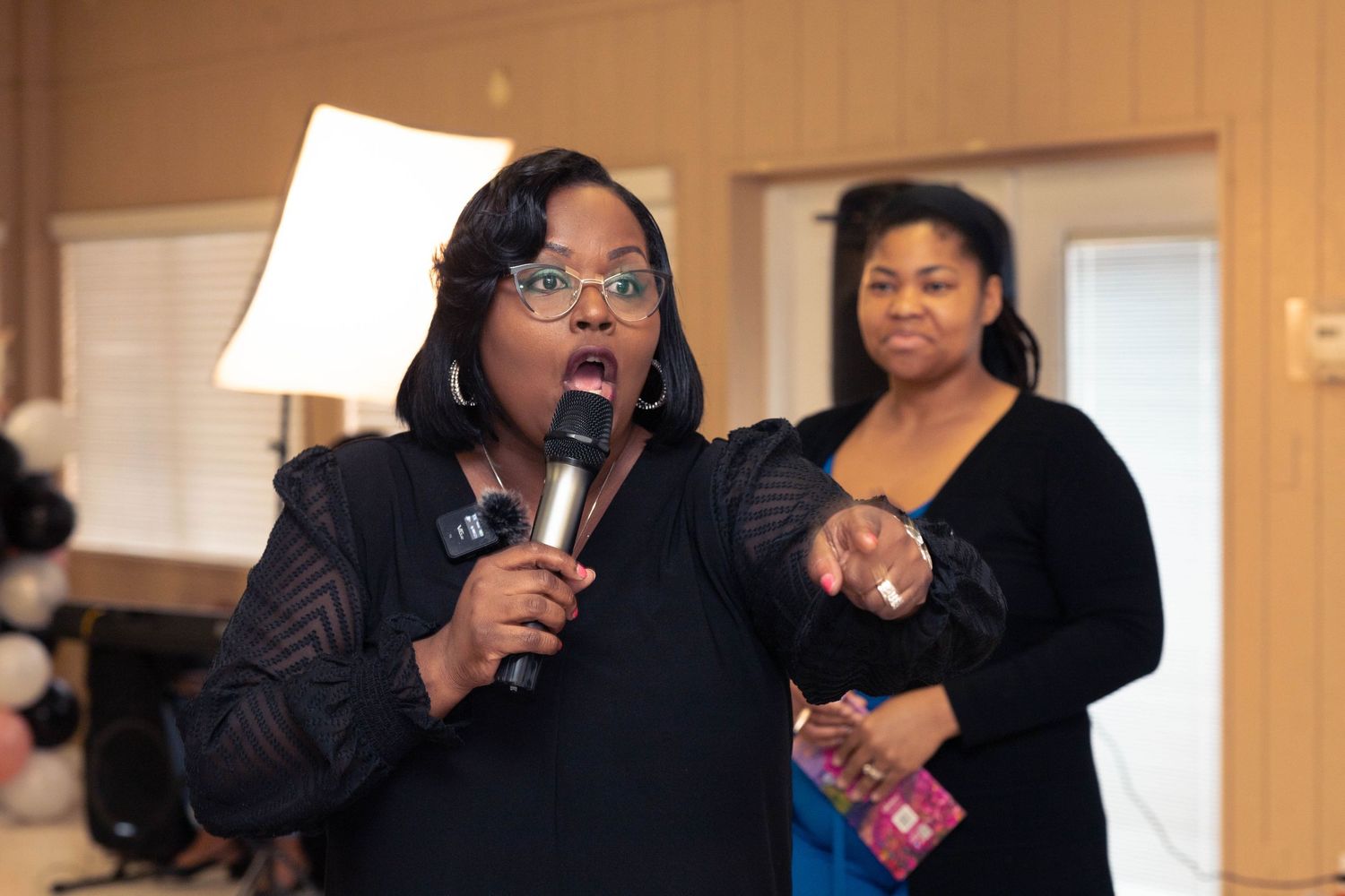Black woman speaking into microphone at indoor event with another woman in background.