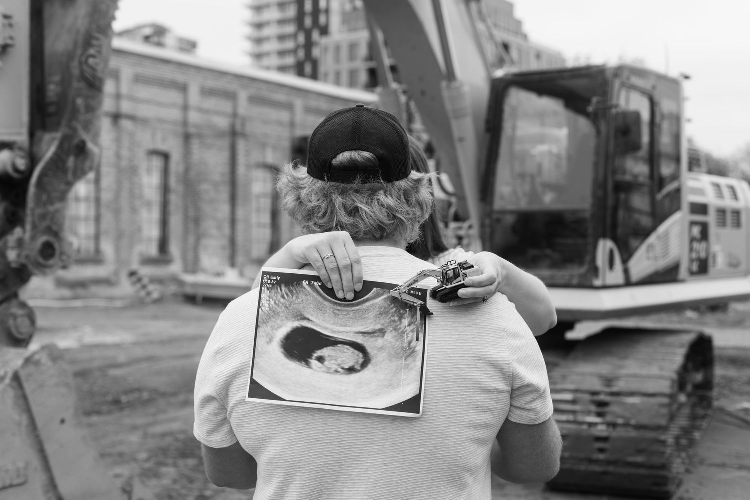 Black and white image showing someone holding an ultrasound photo in front of construction equipment.